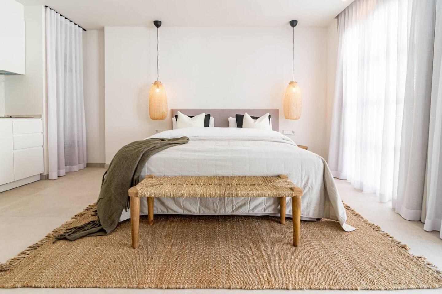 A bedroom with a bed, bedside lamps, a bench, and a large woven rug.