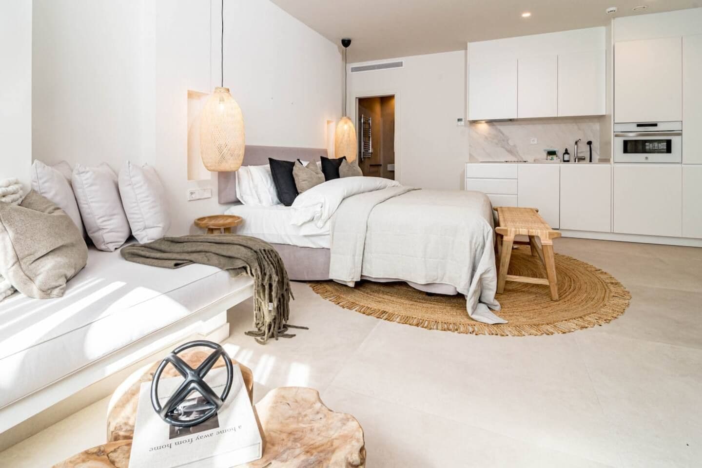 A modern bedroom with a bed, bedside tables, a wooden bench, and a kitchen area with cabinets and appliances.