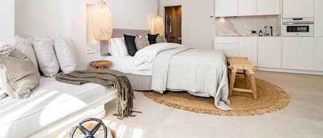 A modern bedroom with a bed, bedside tables, a wooden bench, and a kitchen area with cabinets and appliances.
