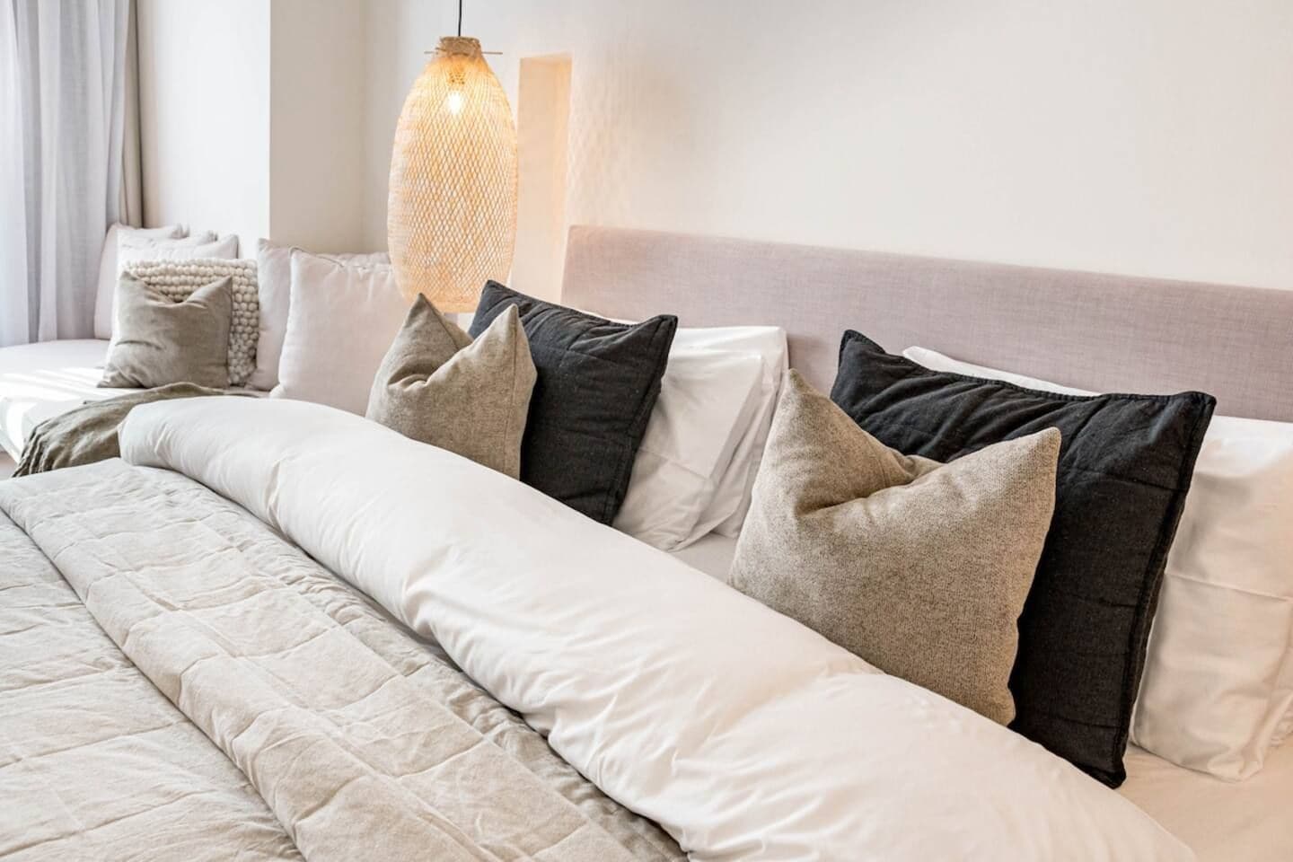 Una cama bien hecha con cubrecama acolchada, varios almohadones en diferentes tonos y una lámpara colgante arriba.