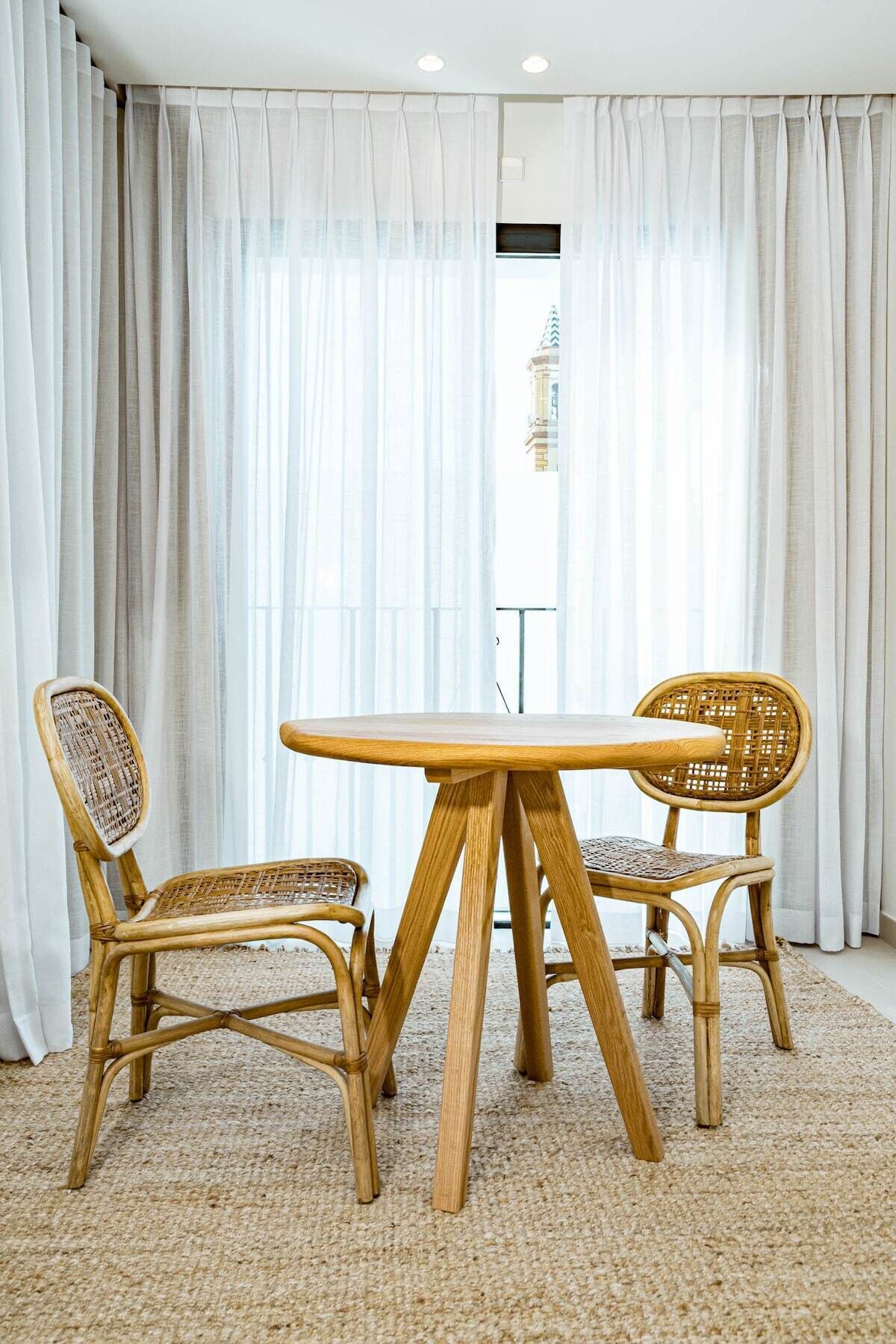 A room with a round wooden table and two wicker chairs, a beige carpet, and sheer white curtains.