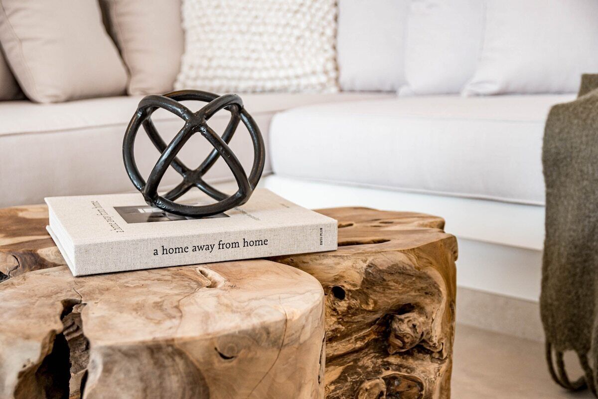 Ein Buch mit dem Titel 'ein Zuhause fernab der Heimat' auf einem hölzernen Hocker, zusammen mit einem gewebten Korb und einer schwarzen Metallskulptur.