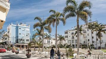 Un área de café al aire libre y soleada, con palmeras, mesas exteriores y edificios al fondo.