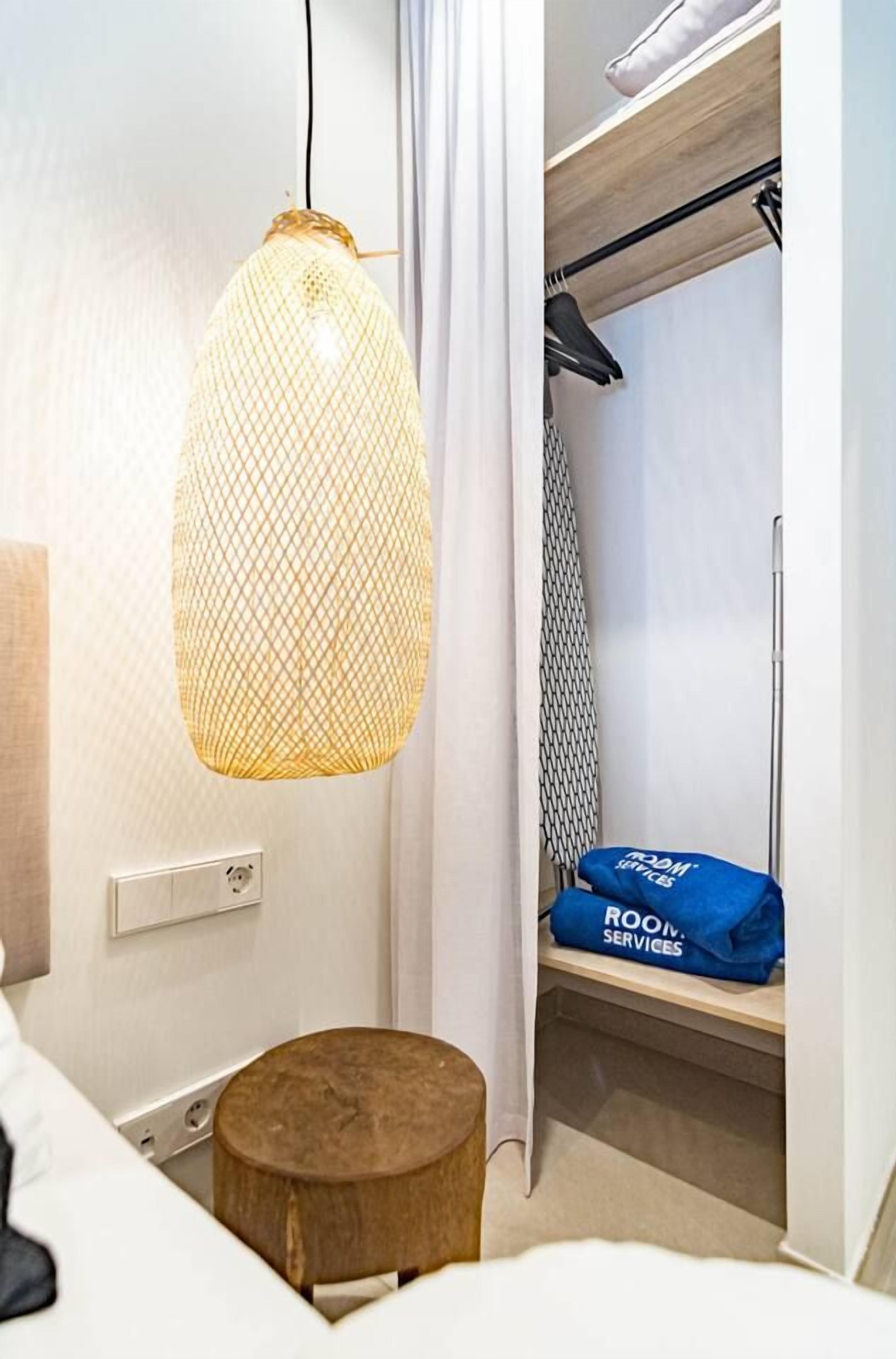 Habitación de hotel con lámpara colgante, un taburete de madera, estante con una bolsa azul y armario con ropa colgada.