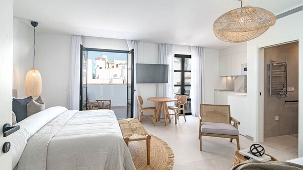 A modern hotel room with a large bed, a dining area, a TV, and a view of the city. - Seaclub Suites by Roomservices (Estepona)
