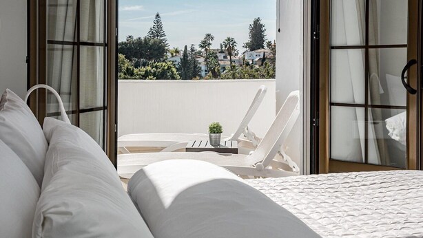 A bedroom with a bed, white pillows, a small potted plant, and a balcony with a chair and table. - AB4 - Lovely Holiday Home Walking Distance, Beach (Marbella)