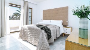 A modern bedroom with a large bed, a wooden headboard, a bedside table with a lamp, and a view of palm trees through a large window. - AB4 - Lovely Holiday Home Walking Distance, Beach (Marbella)