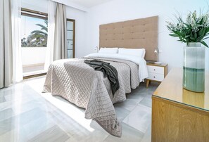 Quarto moderno com uma cama grande, cabeceira de madeira, mesa de cabeceira com abajur e vista para palmeiras através de uma janela ampla.