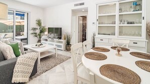 Ein modernes Wohnzimmer mit einer Couch, einem Couchtisch und einem Esstisch mit Platzdeckchen. Es gibt einen Fernseher, eine Vitrine mit Glastüren und einen Topfpflanzen.