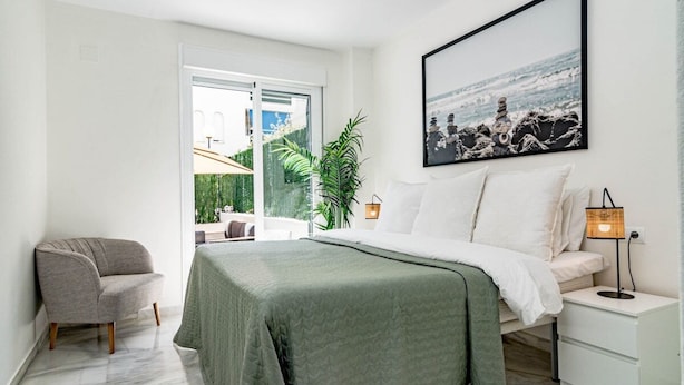 Ein Schlafzimmer mit einem Bett, einem Nachttisch, einem Sessel, einem Fenster mit Blick und einem gerahmten Bild an der Wand.