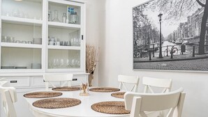 Ein Essbereich mit einem weißen Tisch, gedeckt für sechs Personen, umgeben von weißen Stühlen. Ein großes Fenster mit Einlegeböden zeigt Geschirr und Gläser. An der Wand hängt ein schwarz-weißes Stadtbild.