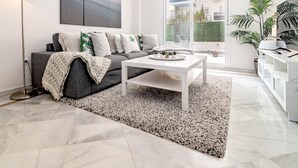 Ein modernes Wohnzimmer mit einem grauen Sofa, einem weißen Couchtisch und einem großen Fenster mit Blick auf Grünflächen.