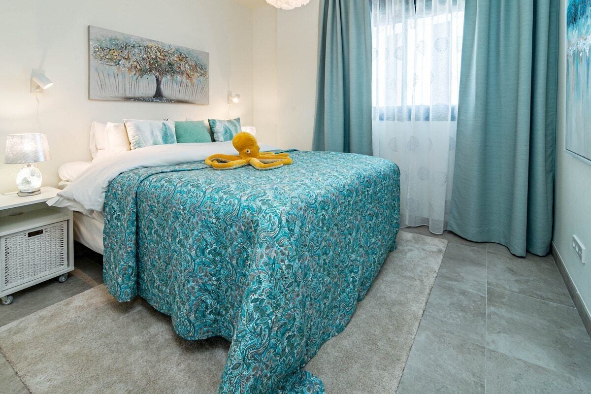 Un dormitorio con una cama, una mesita de noche, una lámpara, un peluche de pulpo y una ventana con cortinas.