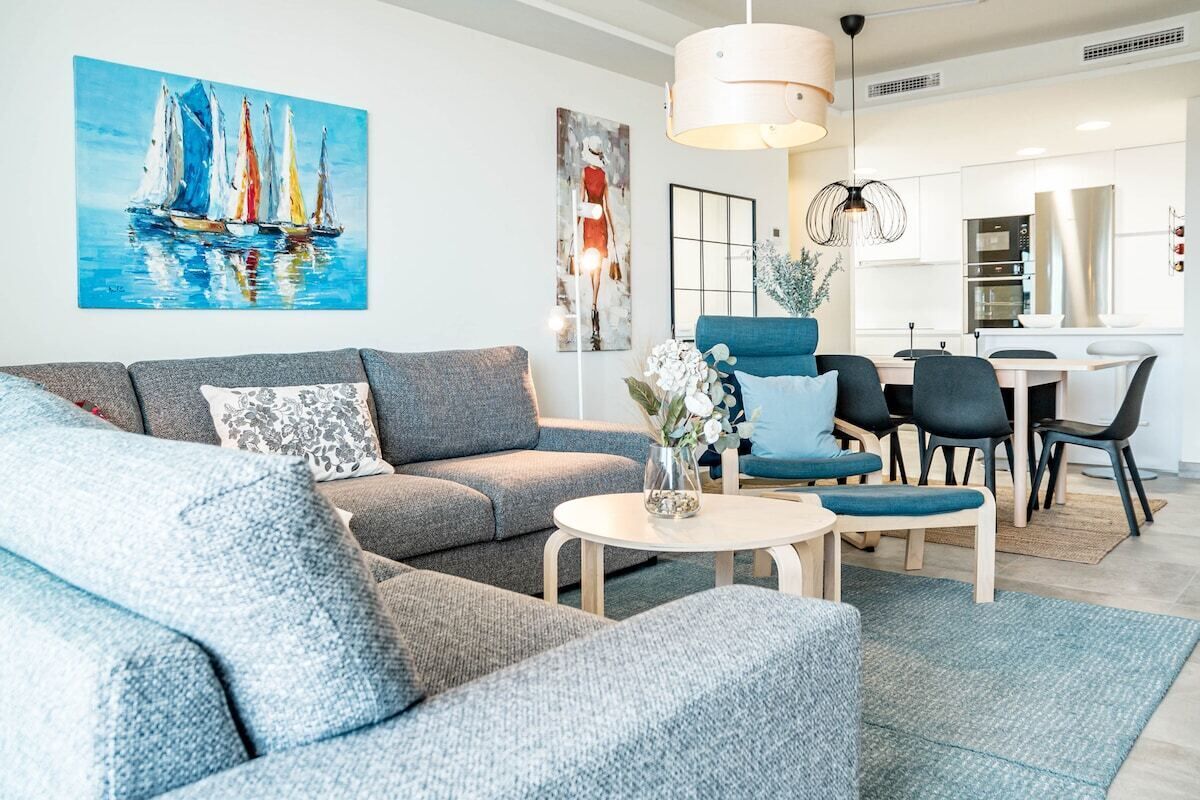 A modern living room with a grey sofa, a round coffee table, and a blue armchair. There is a painting of sailboats on the wall and a kitchen area in the background.