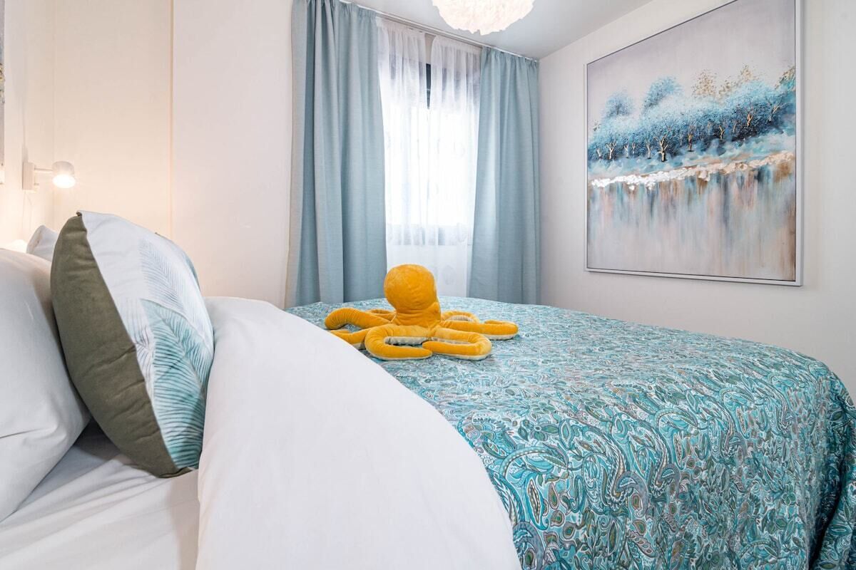 A hotel room with a bed, a large window with curtains, a painting on the wall, and a yellow octopus decoration on the bed.