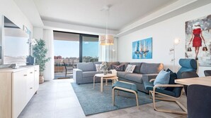 Una sala de estar moderna con un sofá gris, una mesa de centro y un comedor con mesa y sillas. Hay un ventanal que ofrece vistas de la ciudad.