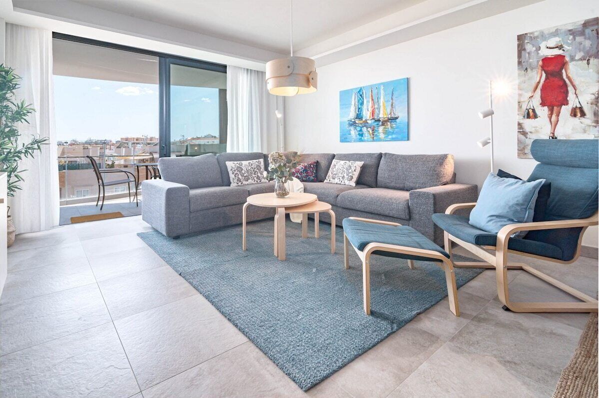 A modern living room with a grey sofa, a blue armchair, and a round coffee table. There is a painting of a sailboat on the wall and a painting of a woman on the floor.