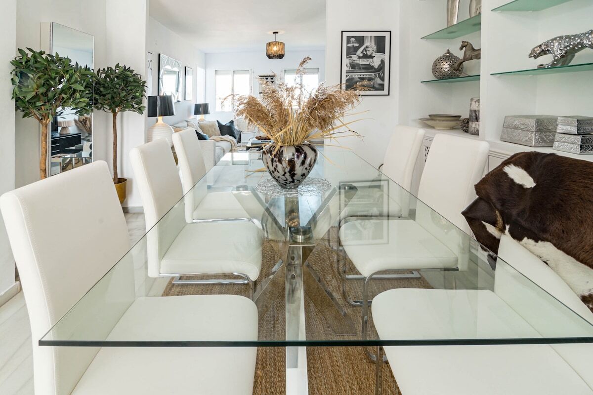 Un espace repas moderne avec une table en verre, des chaises blanches et un vase décoratif contenant des plantes séchées.