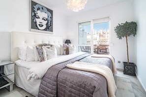 Un dormitorio moderno con una cama grande, una mesita de noche, una lámpara y vistas de la ciudad a través de un ventanal.