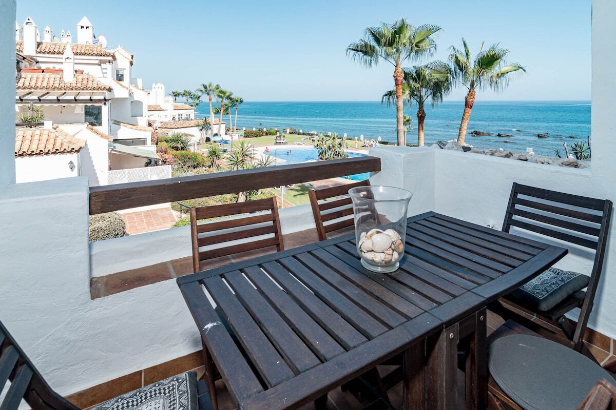 A balcony with a wooden table and chairs, overlooking a pool and beach.