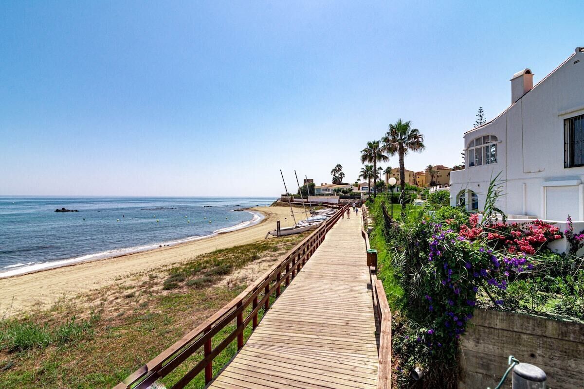 Une promenade en bois menant à une plage où se trouve un bâtiment blanc et des palmiers.