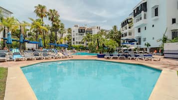 Piscina com espreguiçadeiras e guarda-sóis, rodeada por palmeiras e edifícios modernos.