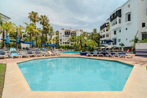 Piscina com espreguiçadeiras e guarda-sóis, rodeada por palmeiras e edifícios modernos.