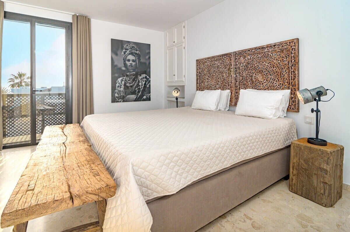 A bedroom with a large bed, a wooden bench, a nightstand, a decorative headboard, and a balcony with a view.
