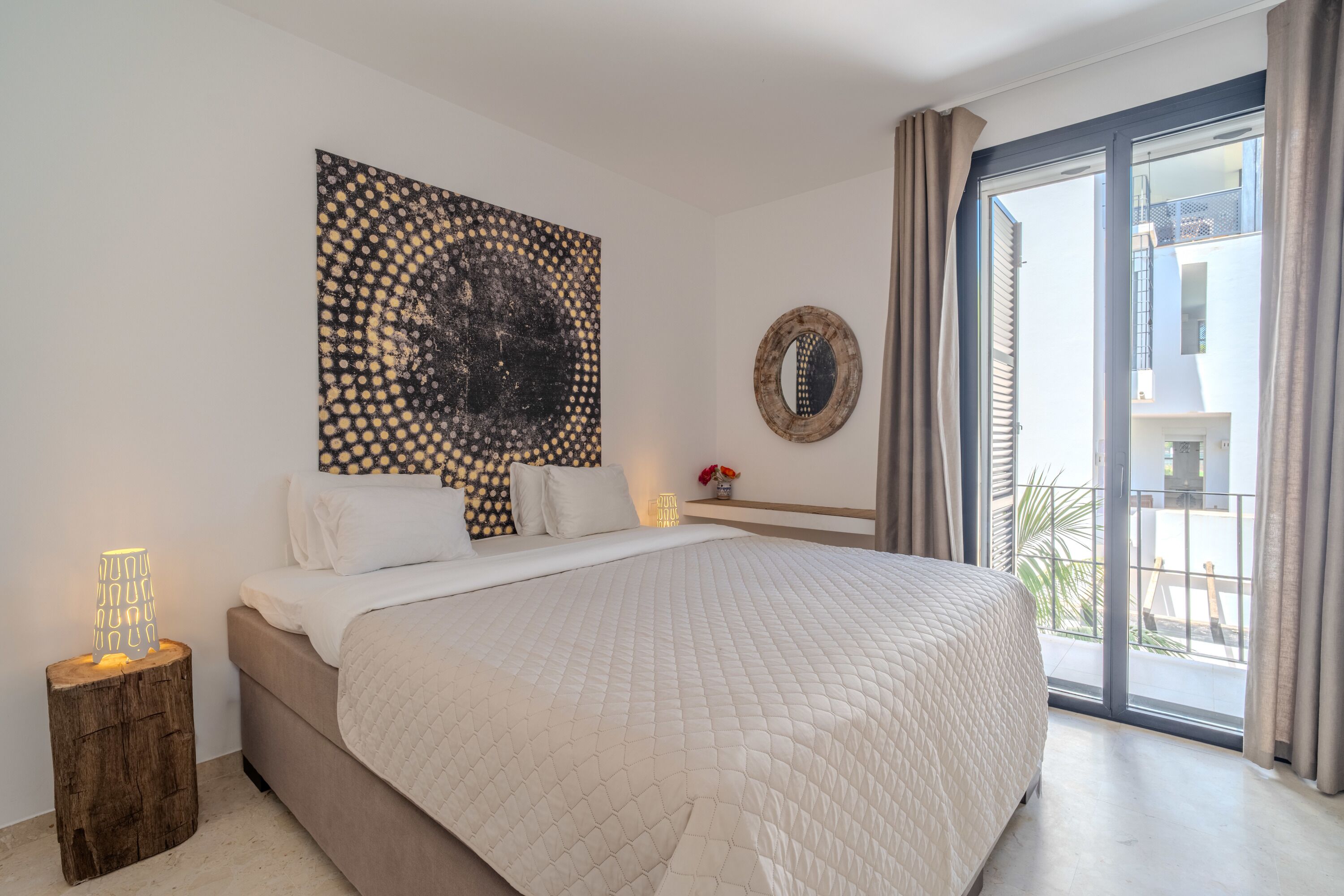 Quarto com cama grande, uma obra de arte na parede, espelho redondo e vista para o exterior.