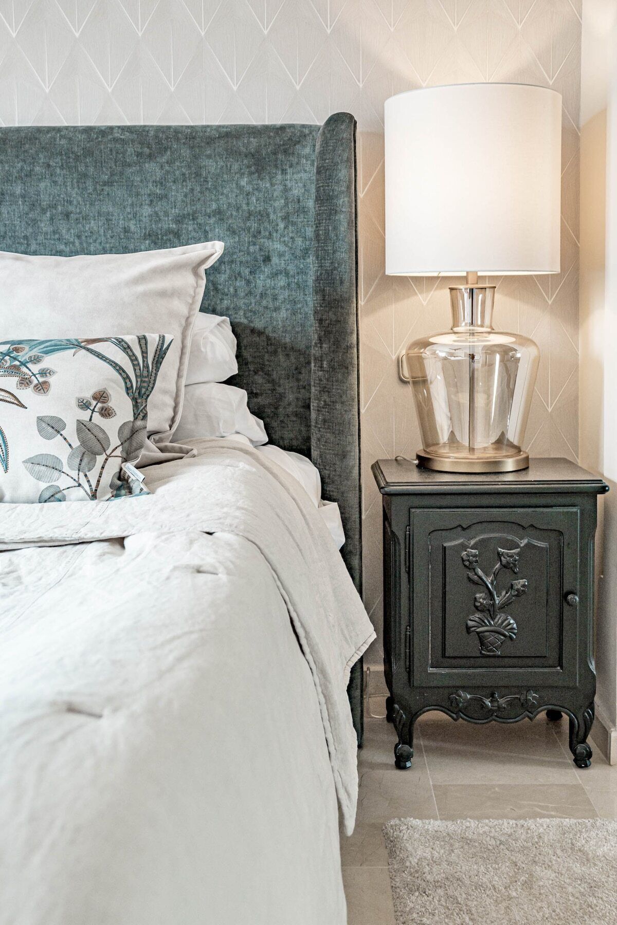 A neatly made bed with a patterned pillow, a dark headboard, a bedside table with a lamp, and a decorative cabinet.