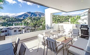 Een modern terras buiten met witte meubels, een barbecue en uitzicht op bergen en groen.