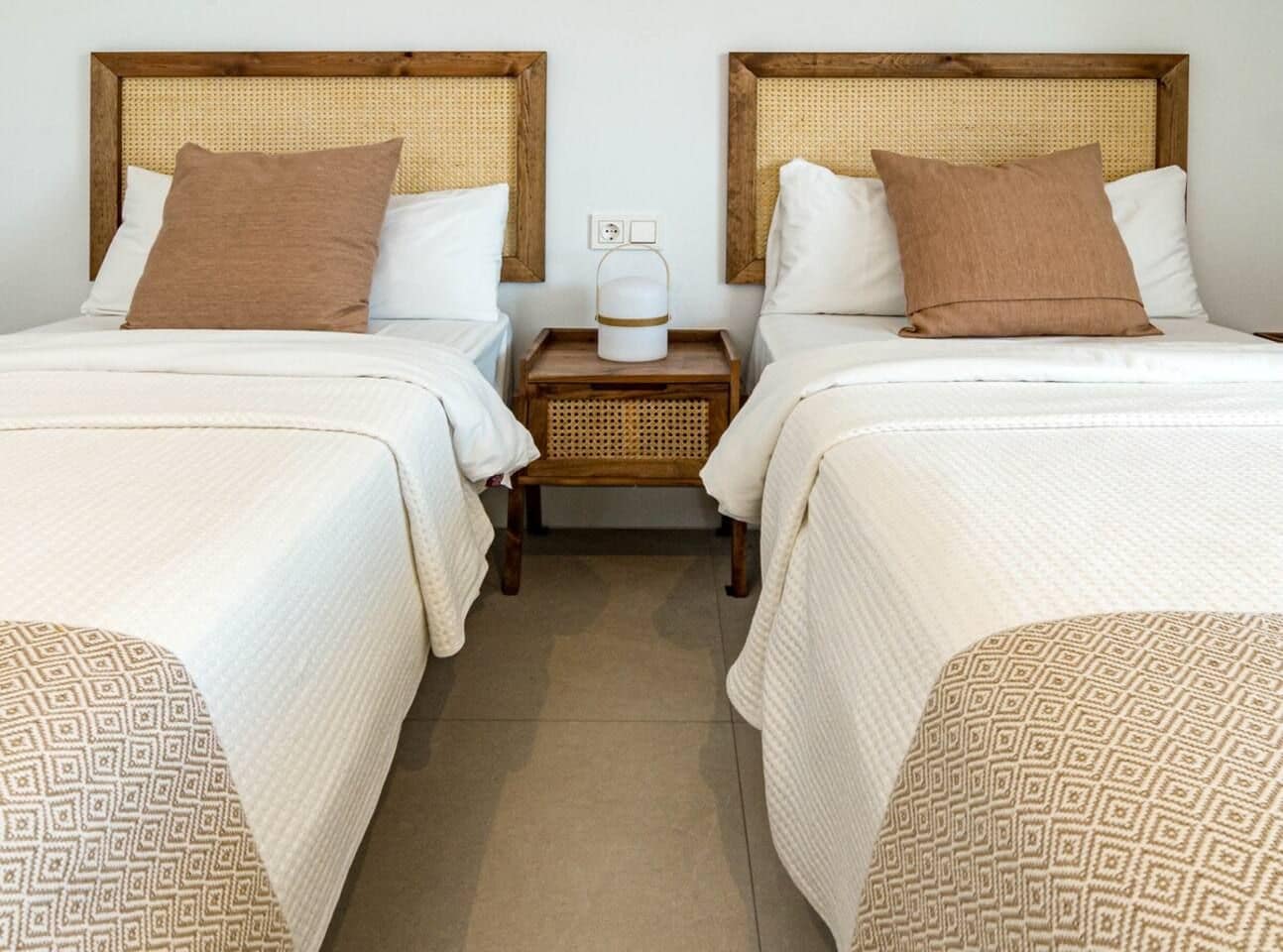 A hotel room with two beds, each with a headboard, a bedside table with a lamp, and a power outlet.