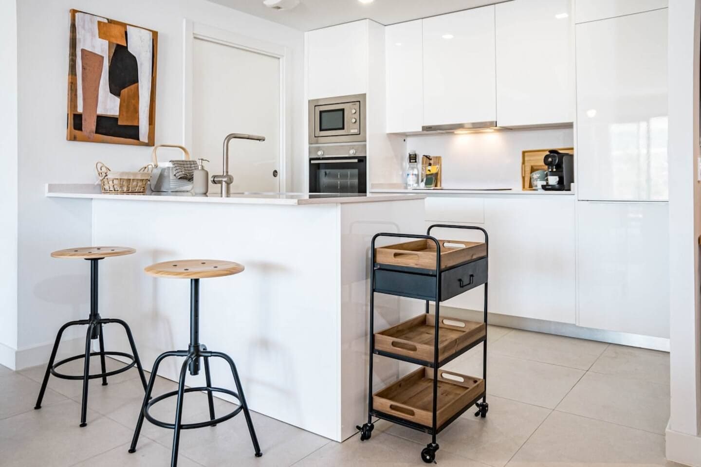 A modern kitchen with white cabinetry, a built-in oven, a microwave, and a coffee machine. There are two stools with round wooden tops and black metal legs. A wooden cart with drawers is also visible.