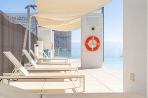 En solbeskinnet udendørs lounge med hvide liggestole og en stor parasol, med udsigt over vandet.