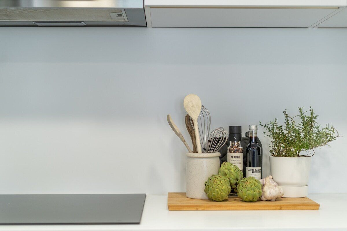 Una encimera de cocina con una tabla de cortar, una taza blanca, una botella de pimienta, una botella de aceite de oliva, una planta de hierbas en maceta y ajos.
