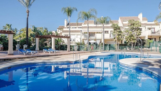 Una piscina con sillones de descanso, palmeras y un edificio con tejado de baldosas.