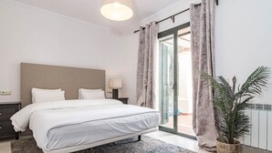 Un dormitorio con cama, mesitas de noche, lámpara, cortinas, una planta y vistas a través de un ventanal.