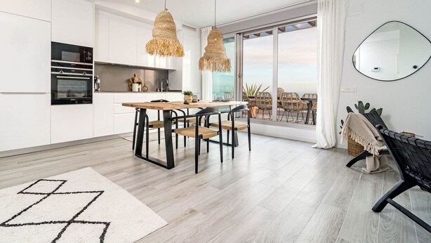Et moderne køkken med hvide skabe, et spiseområde med et træbord og stole, og en stor skydedør, der fører ud til en balkon med udendørs møbler.