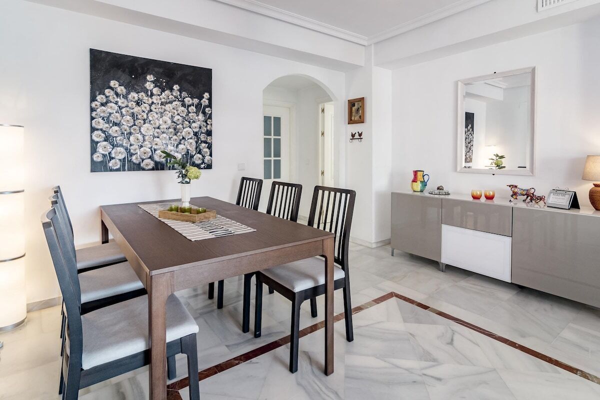 A dining area with a wooden table, six chairs, a vase of flowers, and a painting on the wall.