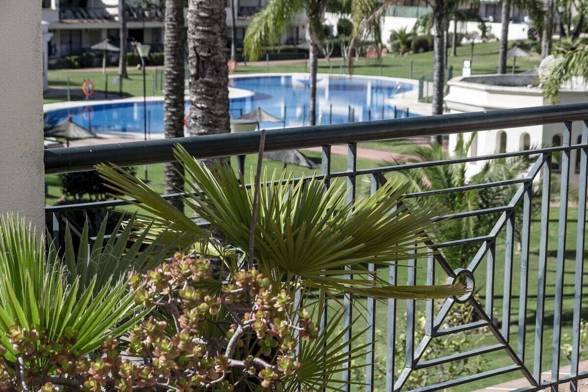 Un balcón con vistas a una piscina, palmeras y un edificio de fachada blanca.