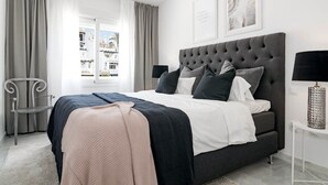 Une chambre à coucher moderne avec un grand lit, des rideaux gris, une vue sur les bâtiments à l’extérieur et un tableau encadré au mur.