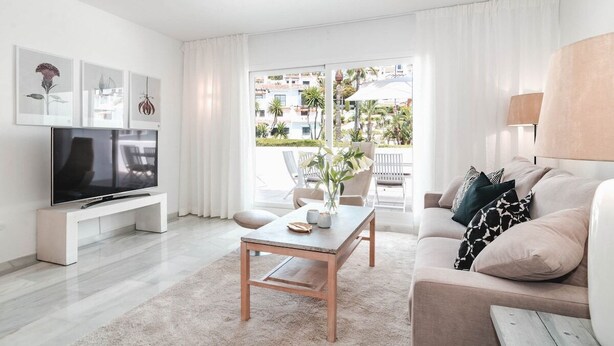 Un salon moderne avec un téléviseur à écran plat, une table basse en bois et un canapé avec des coussins. Il y a un balcon d’où l’on peut admirer les bâtiments et les palmiers.