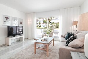 Un salon moderne avec un téléviseur à écran plat, une table basse en bois et un canapé avec des coussins. Il y a un balcon d’où l’on peut admirer les bâtiments et les palmiers.