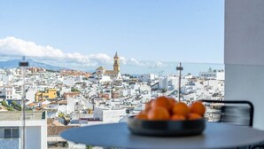 Une vue depuis un balcon, avec un saladier rempli d’oranges posé sur une table, donnant sur un paysage urbain avec des bâtiments blancs et une tour imposante.