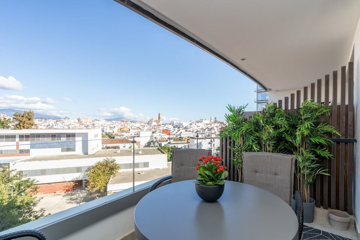Un balcón con vista a un paisaje urbano, una mesa redonda con un florero y plantas en macetas.