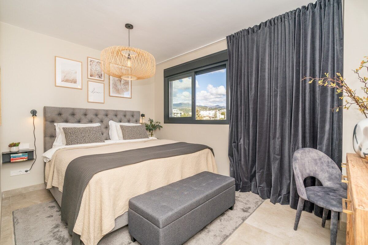 A bedroom with a bed, bedside table, lamp, and a view of the outdoors through a window.