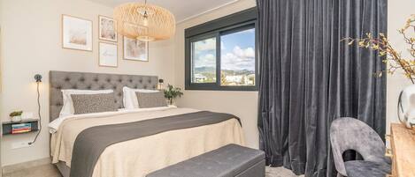 A bedroom with a bed, bedside table, lamp, and a view of the outdoors through a window.