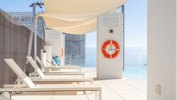 Un espace de détente extérieur, baigné de lumière, avec des chaises longues blanches et un grand parasol, donnant sur une étendue d’eau.
