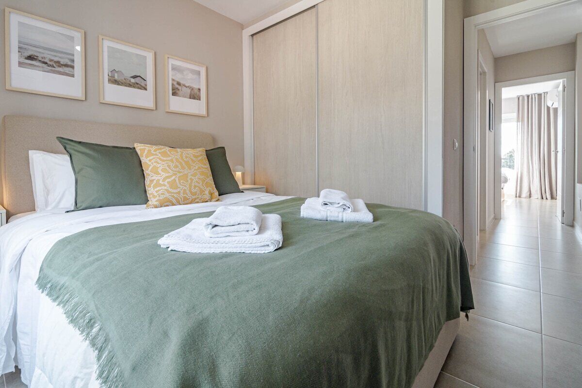 A neatly made bed with a green bedspread, white pillows, and folded towels. There are framed pictures on the wall above the bed.