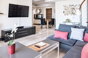Ein modernes Wohnzimmer mit einem grauen Sofa, einem Couchtisch und einer roten Vase mit weißen Blumen. An der Wand ist ein Flachbildfernseher angebracht, es gibt einen Essbereich mit Tisch und Stühlen sowie eine Treppe mit einem außergewöhnlichen Metalldesign.